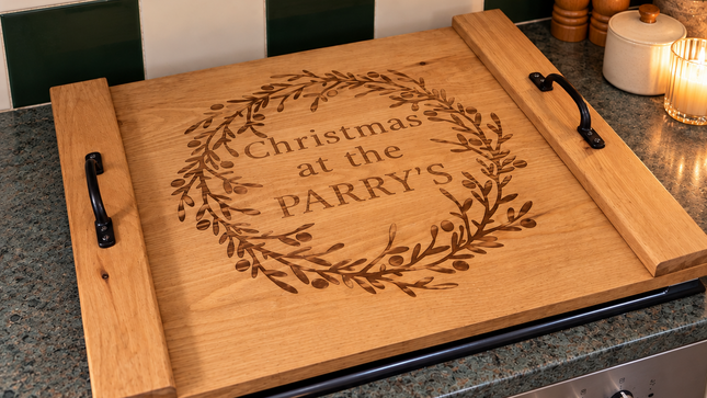A wooden kitchen oven / stove board with black handles, engraved with 'Christmas at the PARRY'S' inside a wreath, resting on a granite countertop in a kitchen setting.