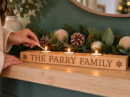 A wooden Christmas mantel candle holder engraved with 'THE PARRY FAMILY' and snowflake details, holding three lit tea light candles on a fireplace mantel with festive greenery and ornaments. A person's hands are shown lighting one of the candles.