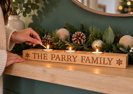 A wooden Christmas mantel candle holder engraved with 'THE PARRY FAMILY' and snowflake details, holding three lit tea light candles on a fireplace mantel with festive greenery and ornaments. A person's hands are shown lighting one of the candles.